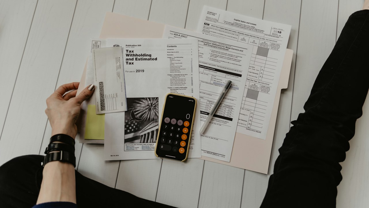 A person at a desk working through printed terms with a smartphone, a pen and a calculator beside the papers