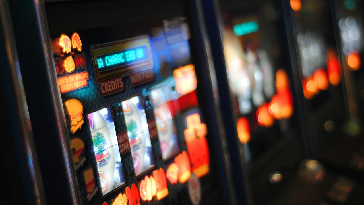 Rows of slot machine screens glowing in a dimly lit casino hall, showing spinning reels and orange status lights